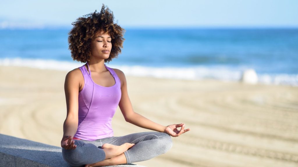 Une femme pratique de la méditation près d'une plage, le vent dans les cheveux et l'air reposé.