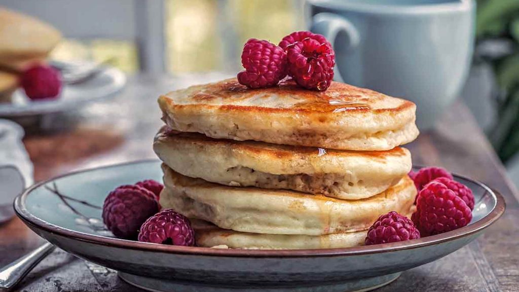 Quatre crêpes véganes épaisses accompagnées de framboises et de sirop d'érable sont déposées dans une assiette bleue.